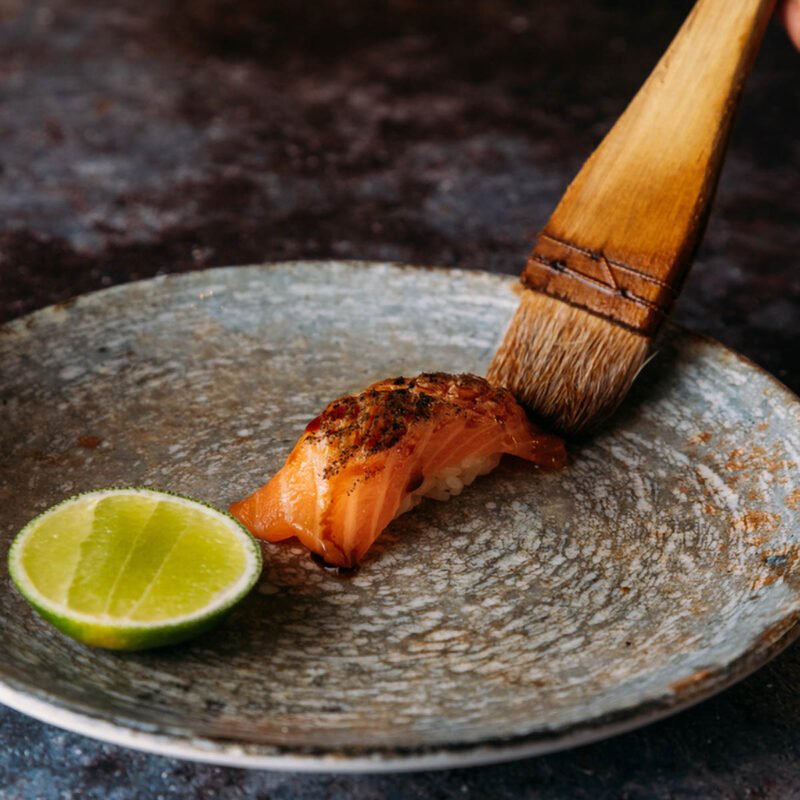A brush finishing a salmon bite with glaze beside a wedge of lime